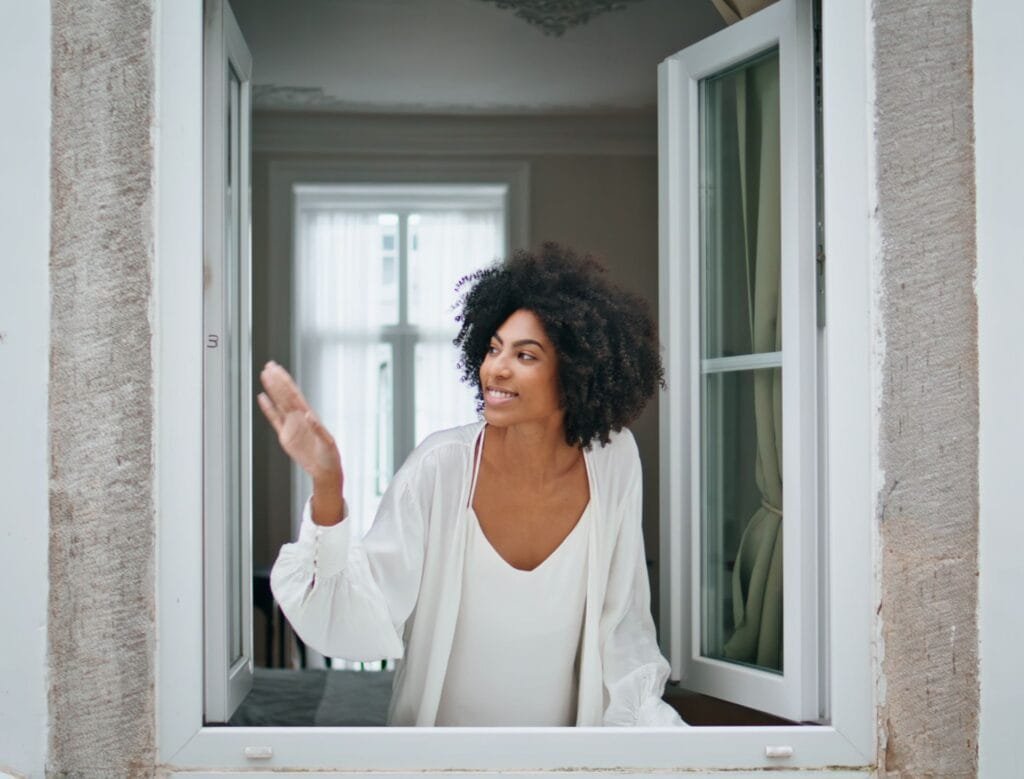 Black hair girl waving window at morning house. Curly smiling woman looking at weather peeping outside. Happy african american model watching cityscape enjoying dreaming at home. Daily routine concept
