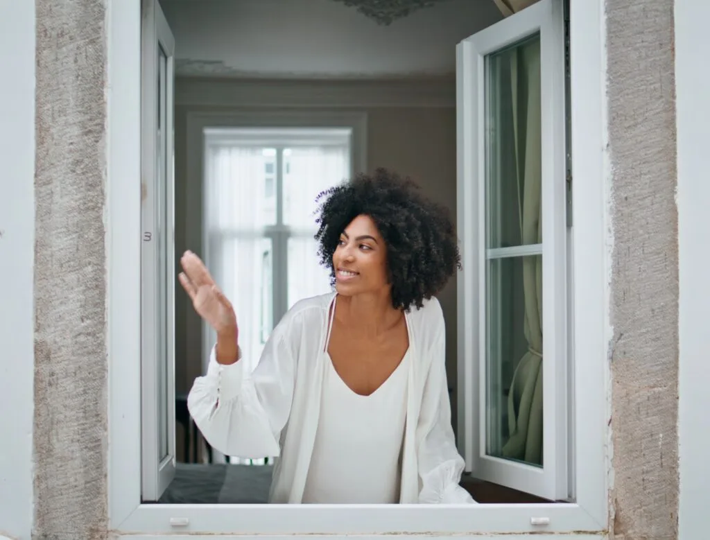 Black hair girl waving window at morning house. Curly smiling woman looking at weather peeping outside. Happy african american model watching cityscape enjoying dreaming at home. Daily routine concept