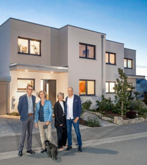 Zwei Familien mit jeweils zwei Paaren und schwarzem Hund stehen vor ihrem gemeinsamen Doppelhaus in der Abenddämmerung.