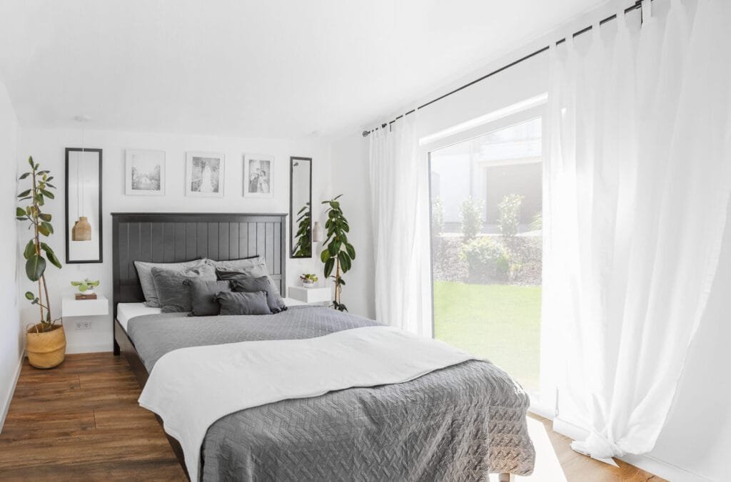 Modernes Schlafzimmer mit Doppelbett und grauer Bettwäsche, weiße Wände und ein großes bodentiefes Fenster mit weißen Gardinen.