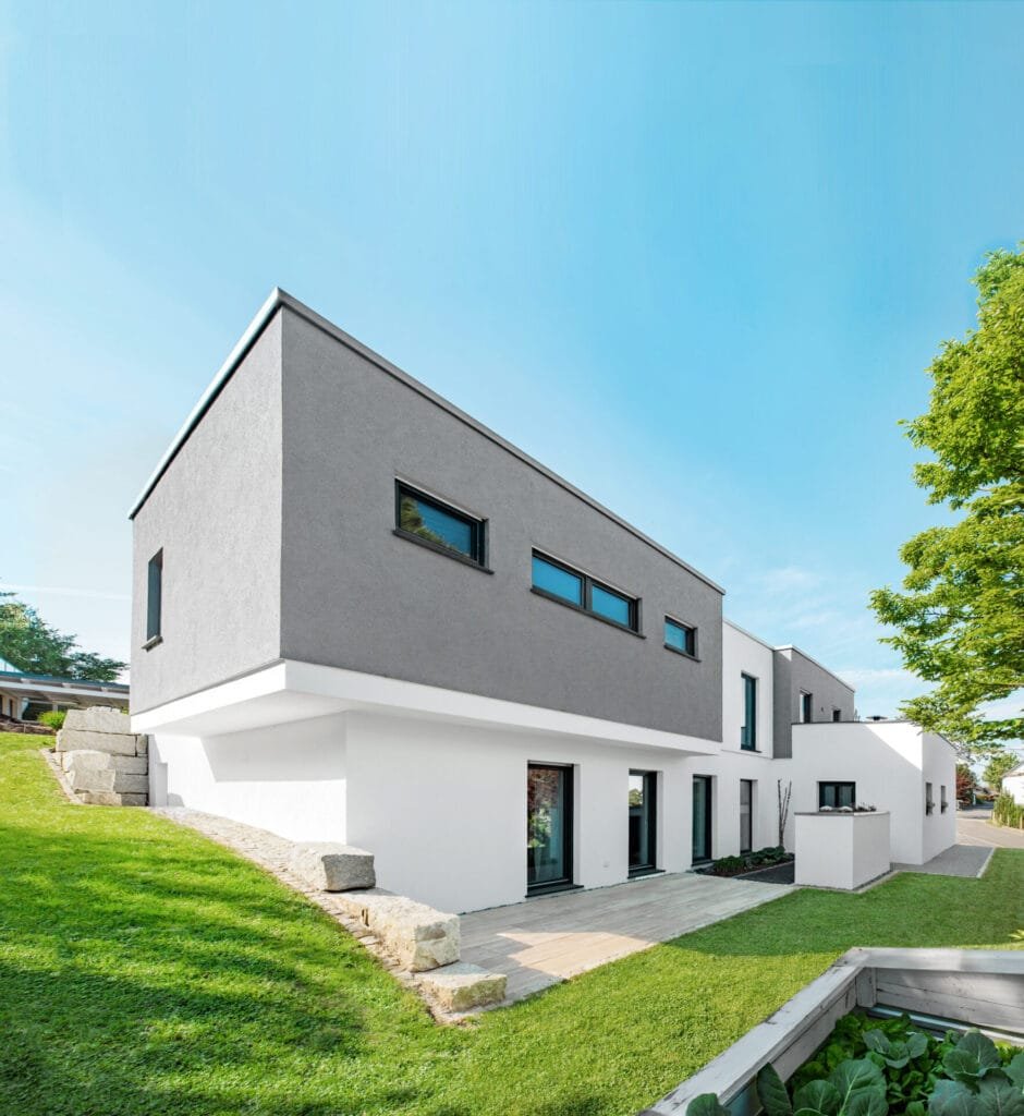 Moderner grau-weißer Bungalow umgeben von einem grünen Garten unter blauem Himmel bei Sonnenschein.