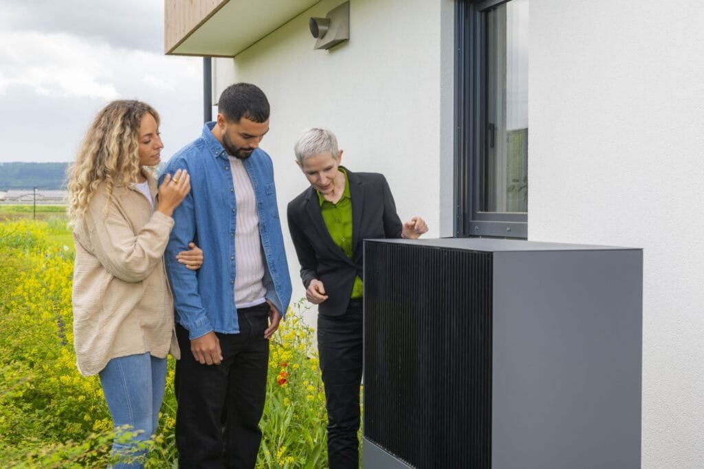 Junges Paar mit Energieberaterin betrachten Außengerät der Wärmepumpe vor einem Haus.