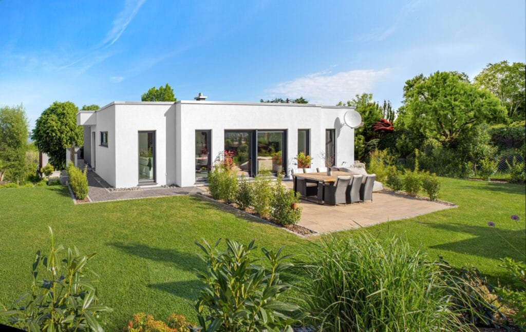 Gartenansicht eines weißen Bungalows am Hang umgeben von einem grünen Garten mit Terrasse unter blauem Himmel bei Sonnenschein.