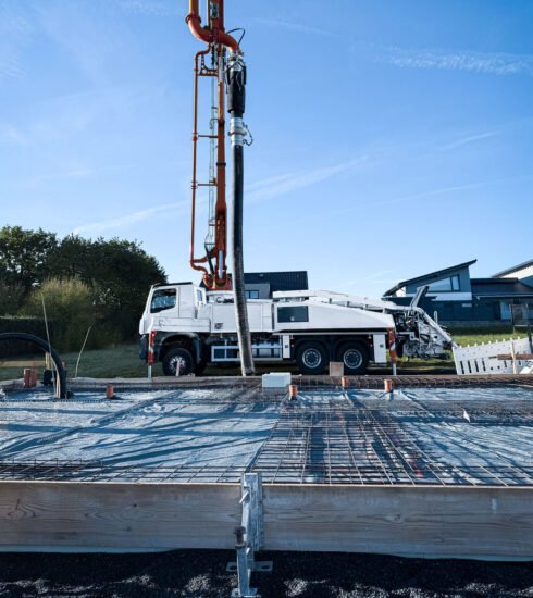 Eine Bodenplatte für ein Einfamilienhaus im Vordergrund wird frisch betoniert. Im Hintergrund ist der Betonlaster mit hohem Kranarm zu sehen der den Betaonschlauch über die Bodenplattenfläche führt.