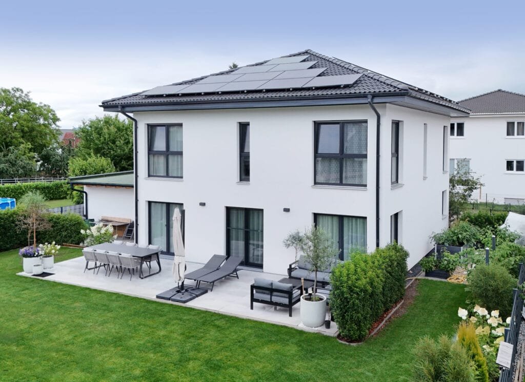 Moderne, weiße Stadtvilla mit großflächiger Terrasse, Gartenmöbeln und Liegen; das dunkle Dach ist mit Photovoltaikmodulen ausgestattet und von grünem Garten umgeben.