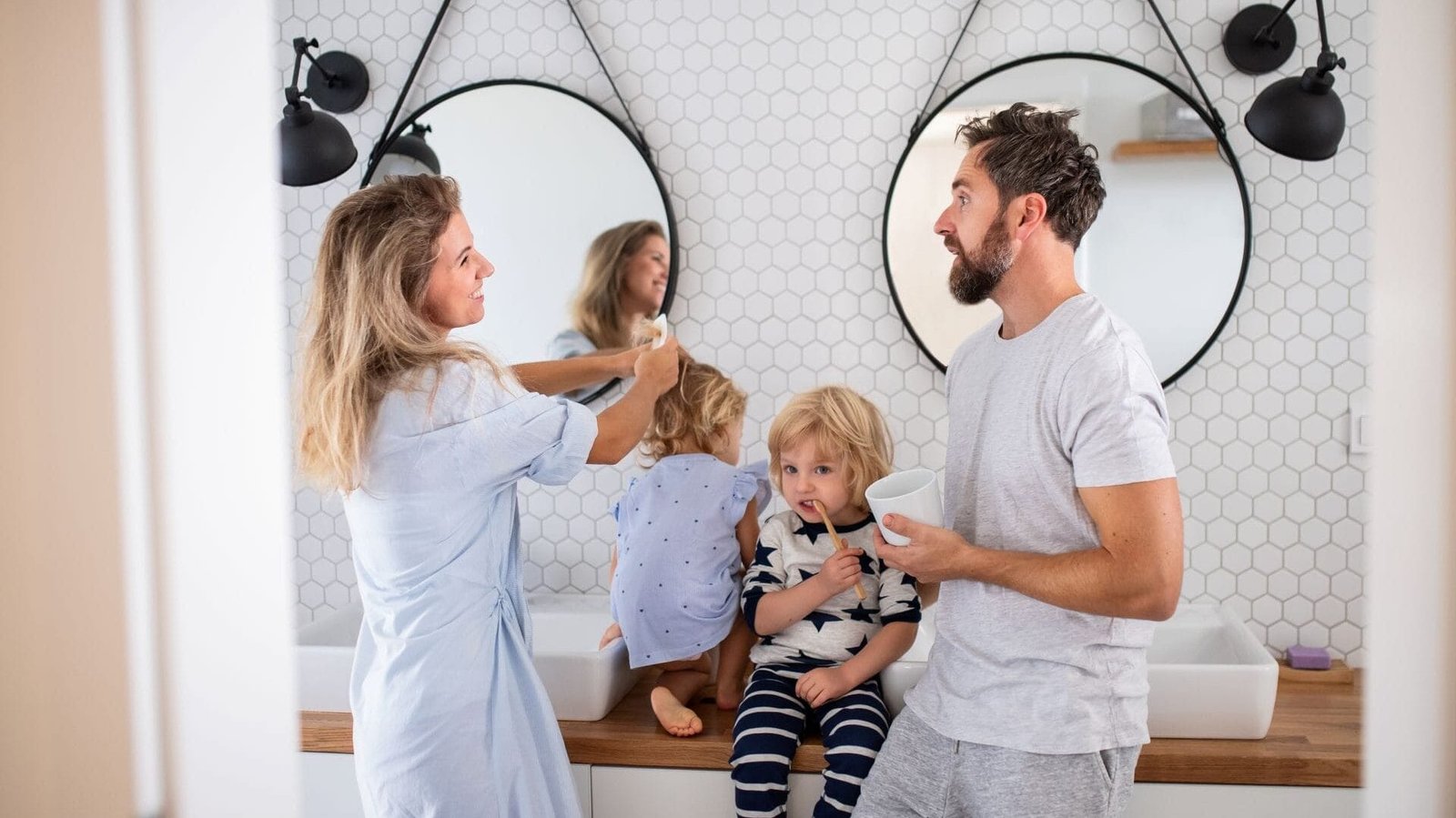 Familie mit zwei kleinen Kindern im Badezimmer. Ein Kind putzt die Zähne, das andere wird von der Mutter gekämmt