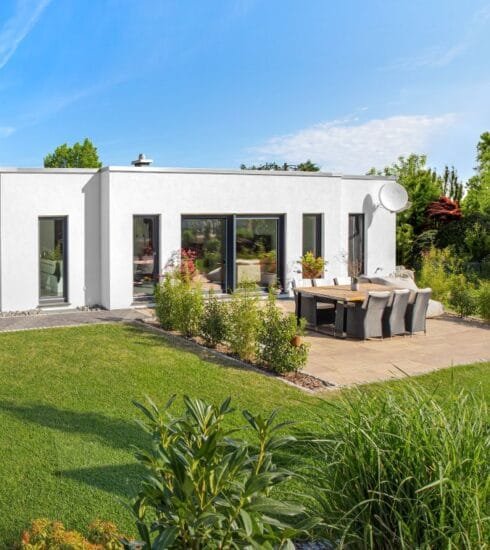 Gartenansicht eines weißen Bungalows am Hang umgeben von einem grünen Garten mit Terrasse unter blauem Himmel bei Sonnenschein.