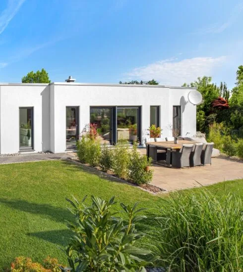 Gartenansicht eines weißen Bungalows am Hang umgeben von einem grünen Garten mit Terrasse unter blauem Himmel bei Sonnenschein.