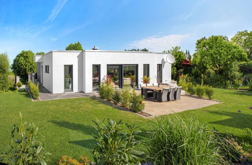 Gartenansicht eines weißen Bungalows am Hang umgeben von einem grünen Garten mit Terrasse unter blauem Himmel bei Sonnenschein.