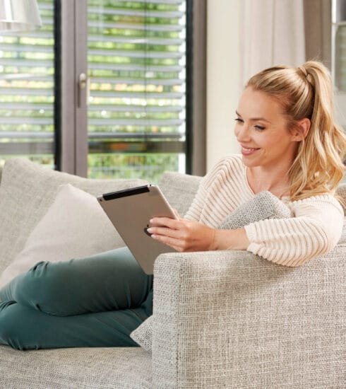 Frau sitzt lächelnd auf dem Sofa in einem hellen Wohnzimmer. Sie hat ein Tablet in der Hand.