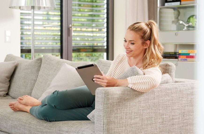 Frau sitzt lächelnd auf dem Sofa in einem hellen Wohnzimmer. Sie hat ein Tablet in der Hand.