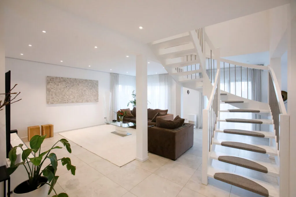 Moderner, offen gestalteter Wohnbereich der Stadtvilla mit eleganter weißer Treppe im Vordergrund, die zu den oberen Etagen führt. Ein gemütliches Sofa und dezente Dekorationen schaffen eine einladende Atmosphäre.