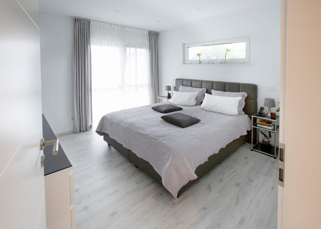 Helles Schlafzimmer mit grauem Doppelbett, hellem Holzboden, bodentiefen Fenstern mit hellgrauen Vorhängen und einem schmalen Querfenster über dem Kopfteil.