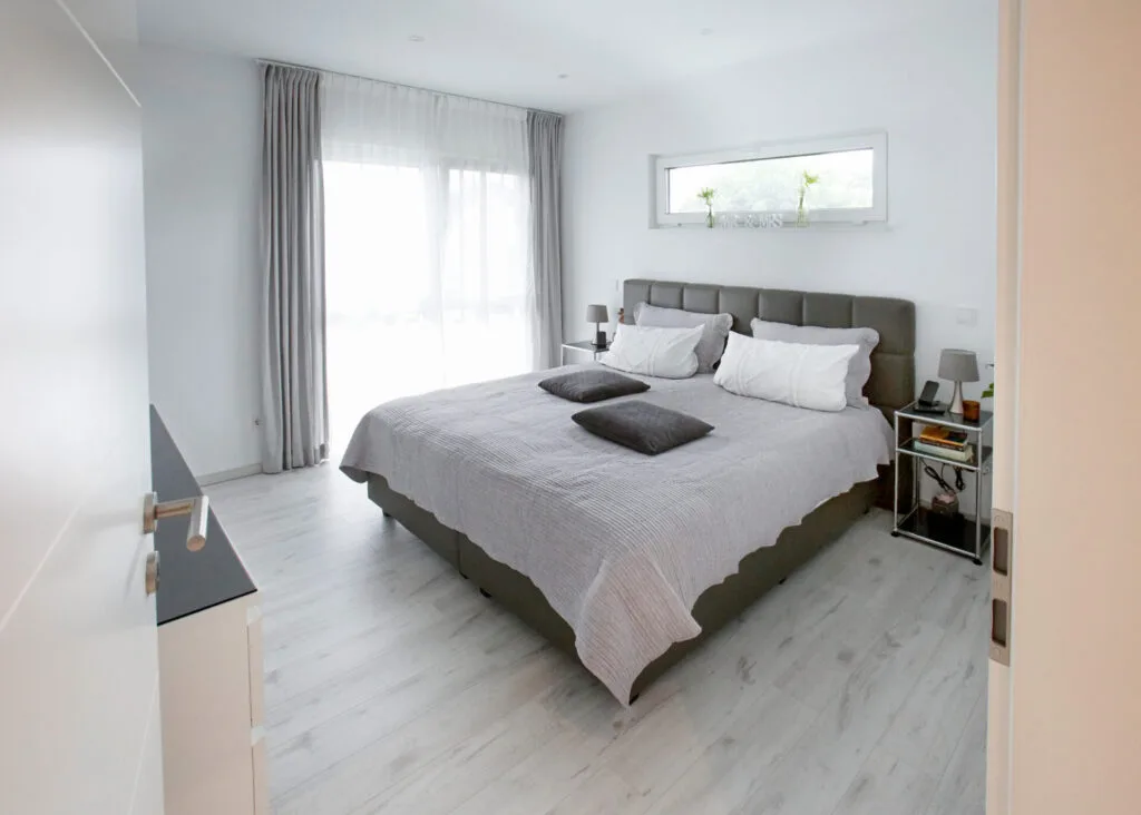Helles Schlafzimmer mit grauem Doppelbett, hellem Holzboden, bodentiefen Fenstern mit hellgrauen Vorhängen und einem schmalen Querfenster über dem Kopfteil.