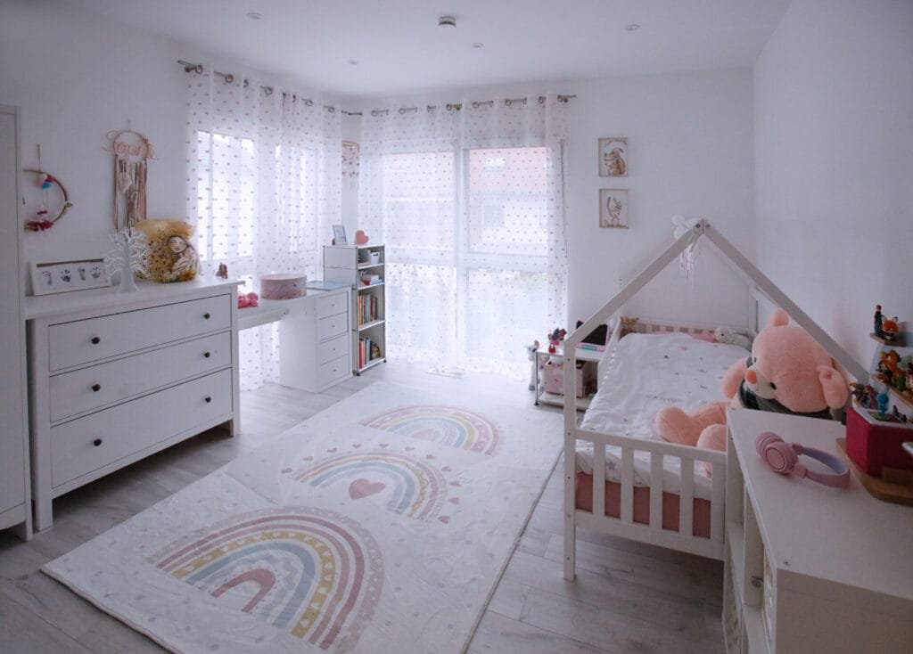 Freundliches Kinderzimmer in Pastelltönen mit weißem Holzhäuschen-Bett, Kommode, Schreibtisch und einem großen Regenbogen-Teppich auf hellem Boden.