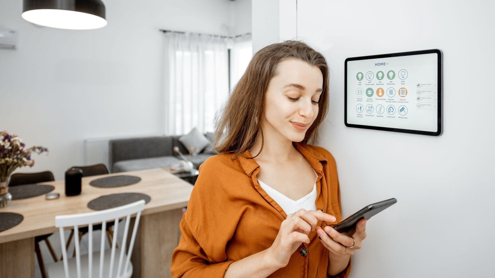 Eine Frau  im Wohnzimmer mit Smartphone in der Hand und tablet an der Wand, das mögliche smarte Steuerungen zeigt.