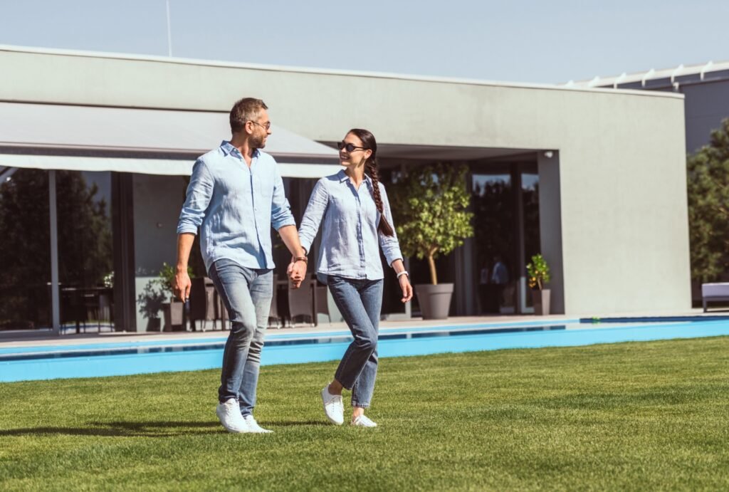 Junges Paar lächelnd, geht Hand in Hand vor modernem Bungalow im Garten