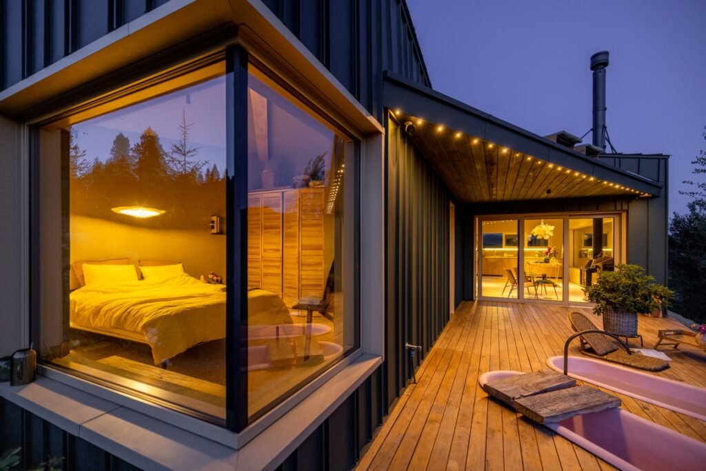 Terrasse eines Bungalows mit Abendstimmung, man kann in das Schlafzimmer schauen sowie weiter hinten am anderen Ende in den Koch-Essbereich des beleuchteten Innenraums.