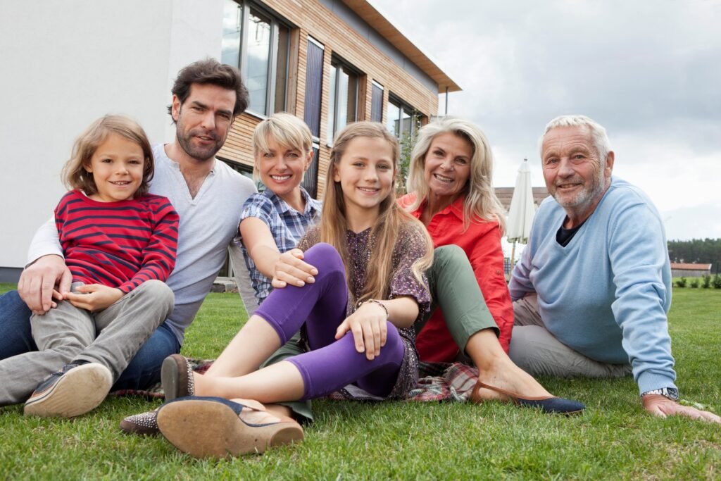 Familie, bestehend aus drei Generationen sitzt im Garten, auf dem Gras, vor ihrem Haus