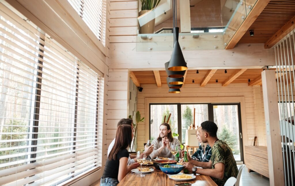 Freunde sitzen am Esstisch, essen und plaudern in einem modernen Block- oder Massivholzhaus mit sichtbaren Holzbalken und -stützen sowie vertäfelten Innenwänden