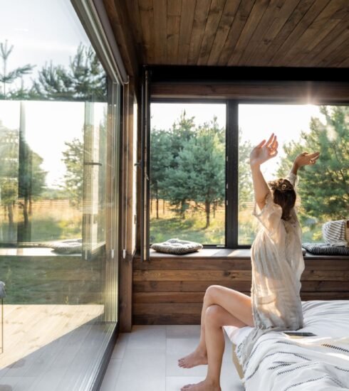 Eine Frau sitzt auf ihrem Bett und schaut durch die großen Übereckfenster hinaus in den lichtdurchfluteten Garten. Foto: RossHelen/www.elements.envato.com