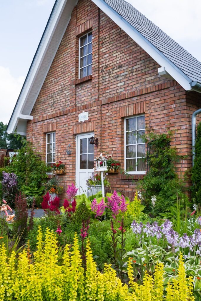 Giebelansicht eines Hauses mit roter Backsteinfassade, weißen Sprossenfenstern und im Vordergrund ein Vorgarten mit bunten Blumen und Stauden