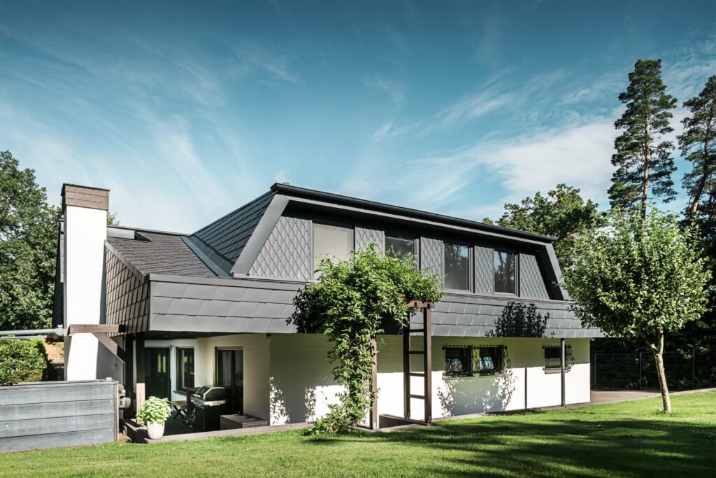 Ein weißes Haus mit grauer Aluminium-Dacheindeckung sowie grauen Rinnen aus Aluminium. Das Haus steht in einem großen Garten mit grüner Wiese und einigen grünen Bäumen.