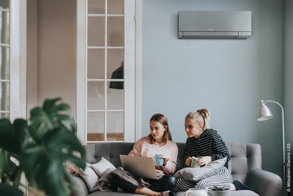 Zwei junge Frauen sitzen auf dem Sofa und lesen in einem Laptop. Hinter ihnen hängt an der Wand eine Klimaanlage von Mitsubishi.