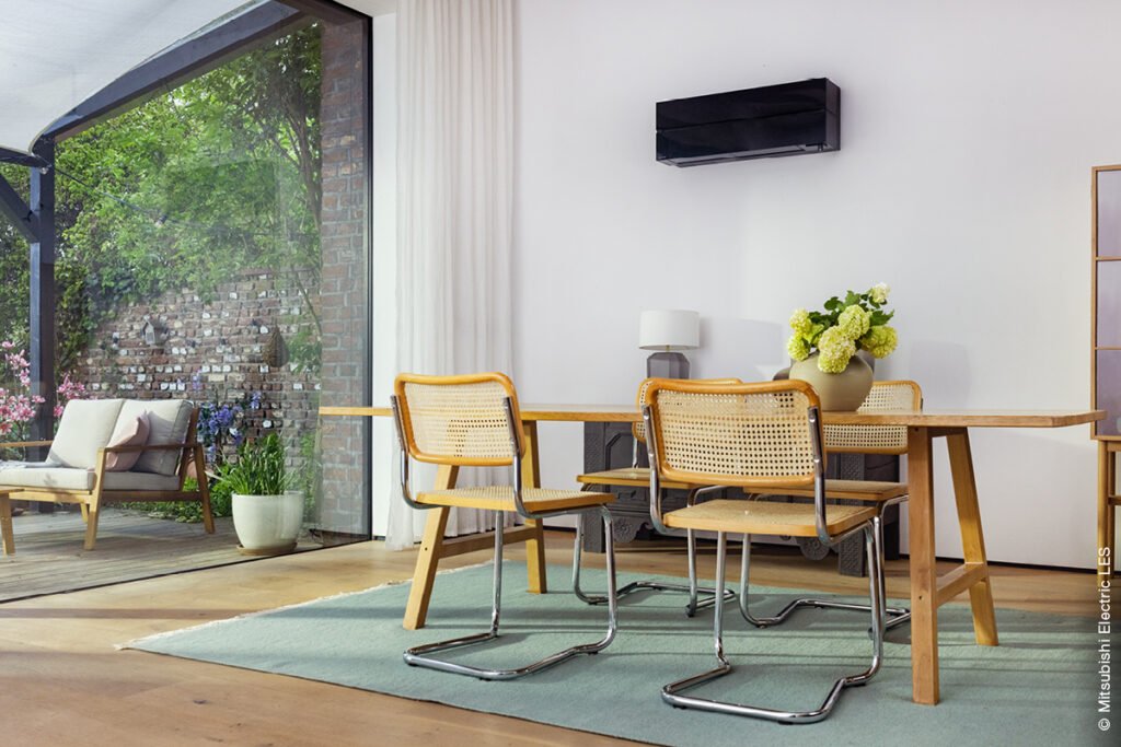 Ein heller Raum mit Essbereich vor einem großen bodentiefen Fenster und Terrasse. An der Wand hängt eine Lüftung von Mitsubishi