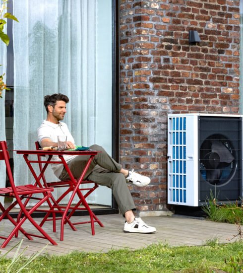 Mann sitzt auf einer Terrasse mit roten Terrassenmöbeln vor einem roten Backsteinhaus. Neben der Terrasse steht eine Wärmepumpe von Mitsubishi Electric.