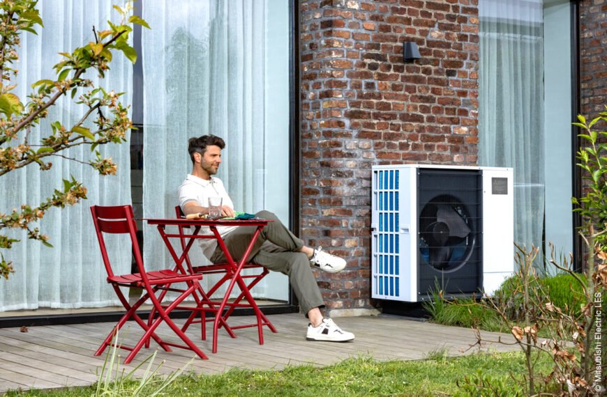 Mann sitzt auf einer Terrasse mit roten Terrassenmöbeln vor einem roten Backsteinhaus. Neben der Terrasse steht eine Wärmepumpe von Mitsubishi Electric.
