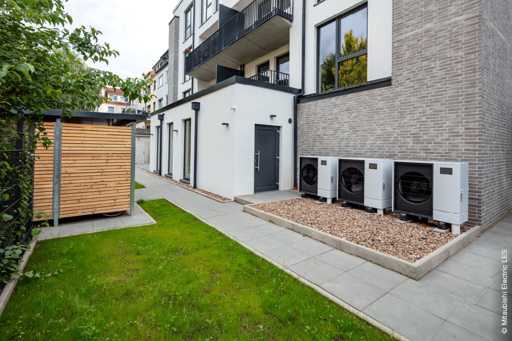 Drei Wärmepumpen, die vor einem Mehrfamilienhaus stehen.