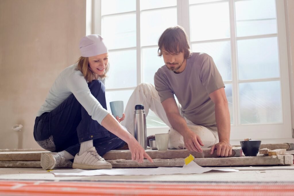 Junges Paar sitzt auf Holzbohlen auf dem Boden und schaut sich Pläne an und trinkt Kaffee - bereit zum Handwerken