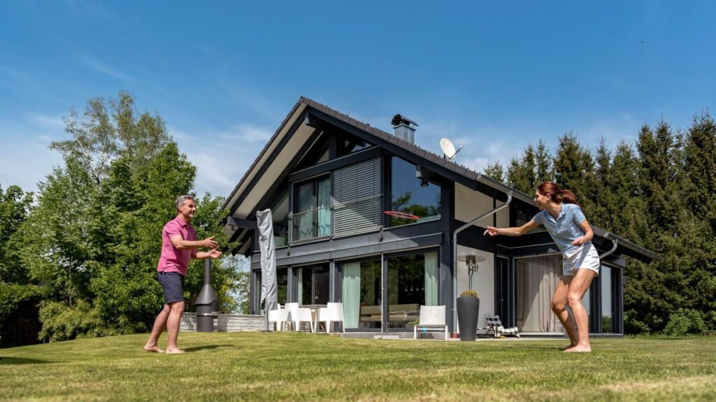 Ein Paar spielt im Garten Frisbee. Das Haus im Hintergrund ist ein modernes Skeletthaus mit vielen raumhohen Verglasungen. 