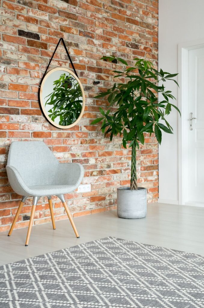 Foto von einem Innenraum mit Spiegel an der Wand, einer hohen Pflanze im Topf, grauem stylischem Stuhl und Ziegelwand