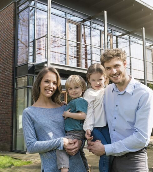 Junge Familie mit zwei Kindern steht lächelnd vor einem modernen Massivhaus
