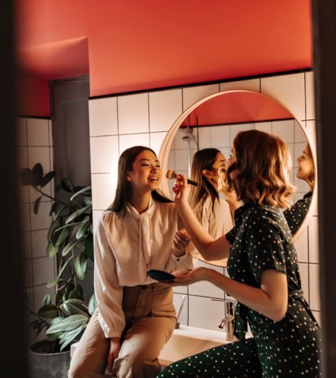Zwei lachende Frauen sitzen auf dem Waschbeckenrand im Badezimmer. Die Frau auf der linken Seite lässt sich von der Frau auf der rechten Seite schminken. Foto: look_studio/www.elements.envato.com