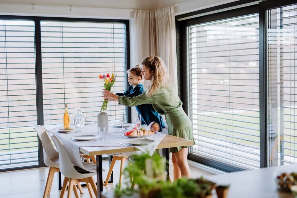 Eine junge Frau stellt mit Ihrer Tochter Tulpen in eine Vase auf einem gedeckten Esstisch vor einer Glasfassade mit heruntergelassenen Raffstores