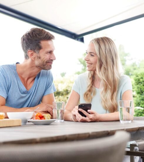 Mann und Frau sitzen auf Terrasse mit Markise und bedienen ein Smartphone