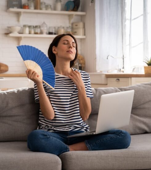 Eine Frau sitzt mit dem Laptop im Schoß auf dem Sofa und fächert sich Luft zu. Foto: Dimaberlin/elements.envato.com